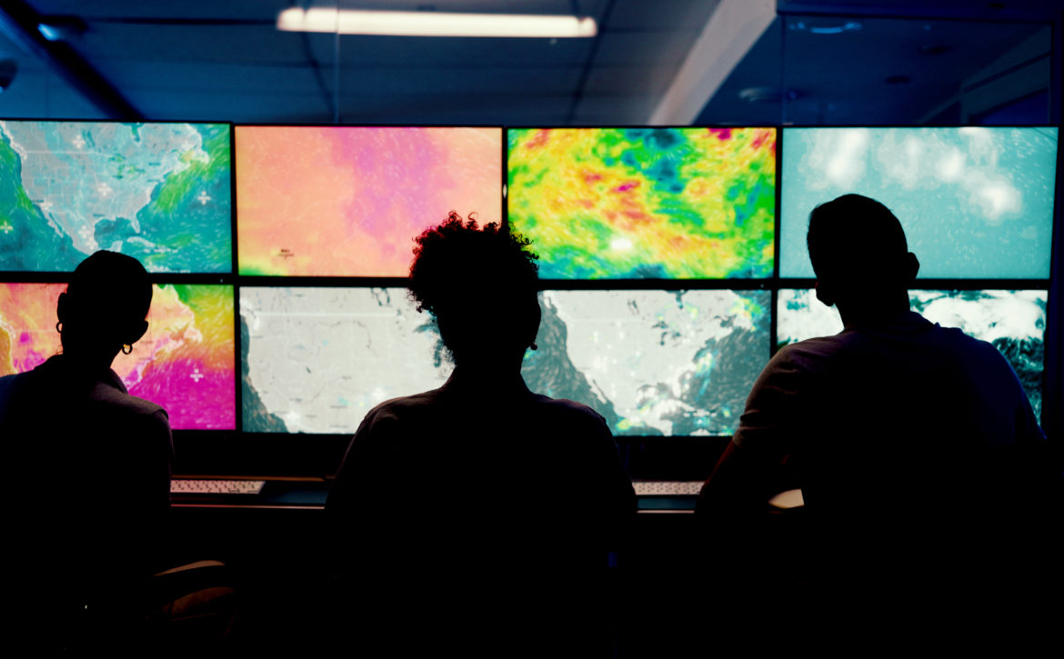 Weather, screen or monitor with people for forecast, satellite maps or teamwork to track storm. Control room, back or scientists with meteorology for broadcast news, report or silhouette at night